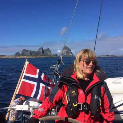 Linda Karlsson på en segelbåt med norska flaggan och höga berg i bakgrunden.