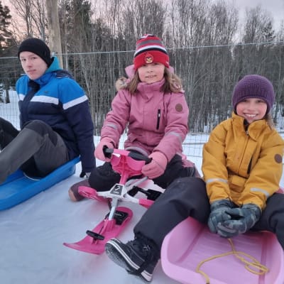 Kolme lasta istuu omilla pulkillaan pulkkamäen yläpäässä.