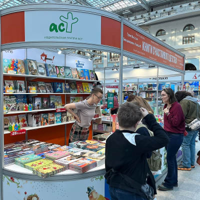 Lastenkirjoja esittelevä koju kirjamessuilla. Myyjä seisoo tiskin takana ja nojaa eteenpäin. Toisella puolella on messujen kävijöitä katsomassa kirjoja.