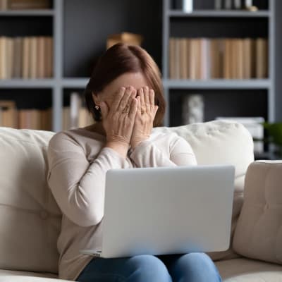 Kvinna sitter i en soffa med datorn i famnen, hon gömmer ansiktet i händerna av skam
