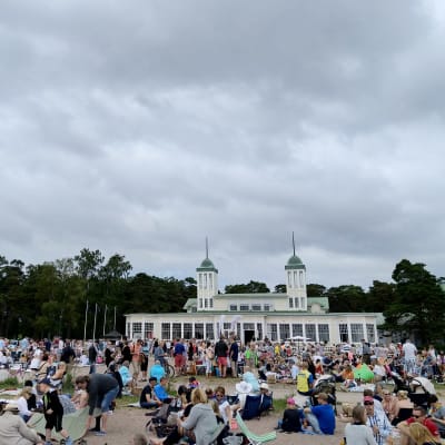 En bild på massor av människor som sitter på en strand och picknickar. I bakgrunden syns ett 1800-tals hus.
