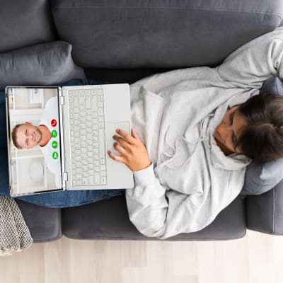 en kvinna ligger i en soffa och har en dator i famnen som hon videochattar med 