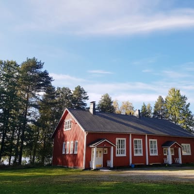 Kuvassa punamultainen vanha kansakoulurakennus kesällä, valkoiset ikkunanpuitteet, taustalla järvi.