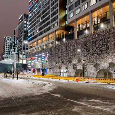 Fasaden på ett köpcentrum, fotat från andra sidan av en väg. Vid fasaden finns röd-gula staket som stoppar fotgängare från att gå på trottoaren. I bakgrunden syns en lyftkran. Det är mörkt ute, och lite snö på marken.