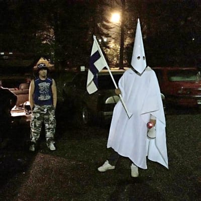 Tre personer under en demonstration mot flyktingar. En person är klädd i Ku Klux Klan-kläder och bär en finsk flagga.