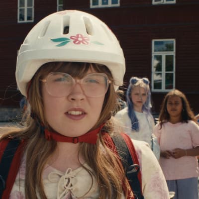 En flicka med glasögon och cykelhjälm tittar med intensiv blick mot ut mot skolgården.