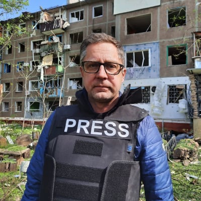 Toimittaja Antti Kuronen tuhoutuneen kerrostalon edessä. Antilla suojaliivi jossa lukee press