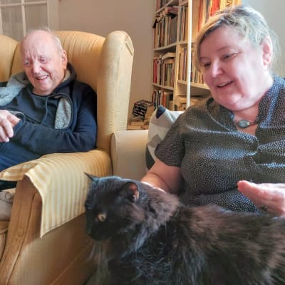 Författaren och diplomaten Lars Bergquist skrattar i sin länstol med författaren och historikern Carina Nynäs sittande bredvid sig i soffan. Också hon ser glad ut med katten Felix i famnen. November 2021.