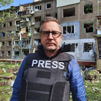 Redaktör Antti Kuronen framför ett sönderskjutet höghus. Antti är klädd i en skyddsväst med texten "Press".
