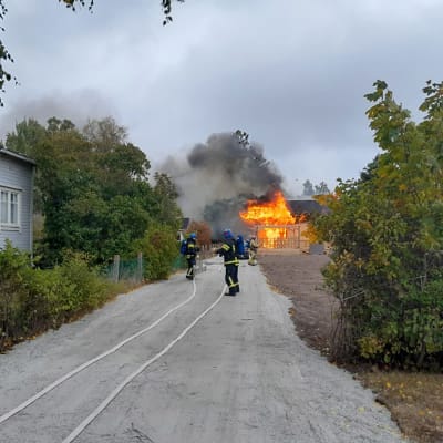 Ett hus brinner i bakgrunden, brandmän på väg att släcka