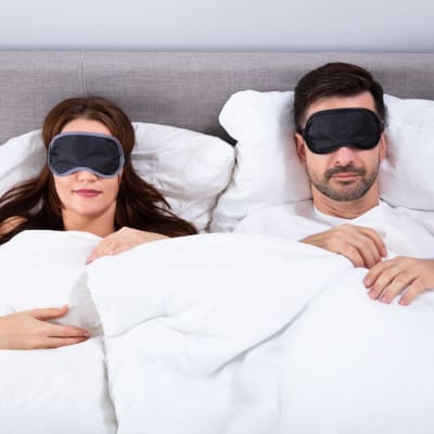 Kvinna och man ligger i en säng, båda har svarta sov-masker över ögonen