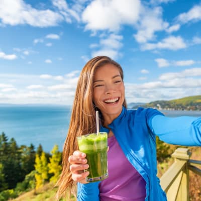 Leende ung kvinna tar en selfie framför en vacker vy och med en grön smoothie i handen