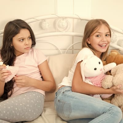 Två småflickor sitter i en säng, den ena ler stort och den andra tittar surt på henne