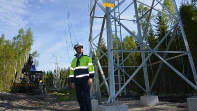 Juha Rintamäki står bredvid en elstolpe i stål i skogen. Han har neongula arbetskläder och hjälm på sig. 