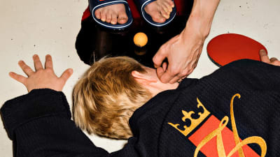 En man i svart morgonrock ligger på golvet med en blodpöl runt ansiktet. En hand sträcker sig ner och rör vid hans kind.