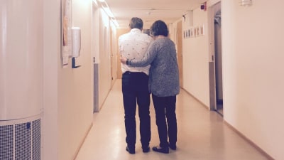Mies ja nainen seisovat selin kameraan päät painuksissa lähekkäin sairaalan käytävällä. Nainen on kiertänyt kätensä miehen ympärille.