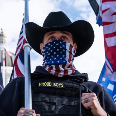 En man i cowboyhatt står framför Capitolium i USA. Han bär en scarf med USA:s flagga över ansiktet, på hans jacka står det "Proud boys".