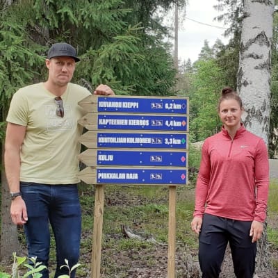 Marko Anttila vaaleankeltaisessa t-paidassa ja Jenni Hiirikoski vaaleanpunaisessa juoksutakissa seisovat uusien opaskylttien vieressä metsän siimeksessä.