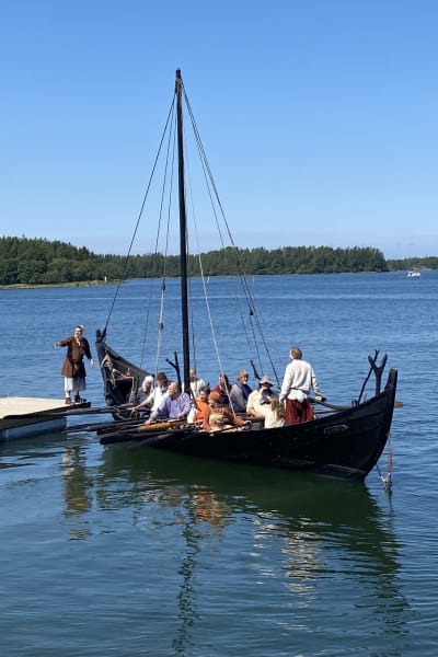 Vikingaskeppet Sotka med ett tiotal roddare lägger till vid en brygga i skärgården.
