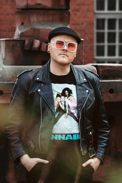 Eino Nurmisto seisoo vanhan tehdasrakennuksen edessä. Hän pitää käsiä housujensa taskussa ja katsoo vakavalla ilmeellä kameraan. Yllään hänellä on mustat vaatteet. Paidassa Spice Girlsin kuva ja teksti Wannabe.