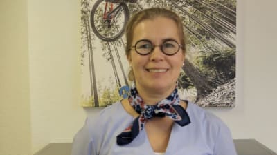 Kvinna i lila skarf och glasögon ser rakt in i kameran och ler. 