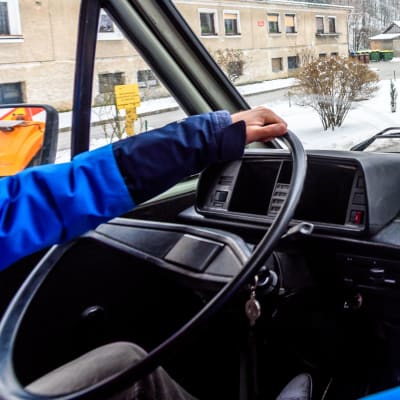 Händer på en ratt i en lastbil och vy över ett vintrigt landskap och väg genom fönsterrutan.