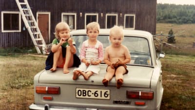Merja Kyllönen istuu pikkulapsena kahden serkkunsa välissä Ladan takakontin päällä. Kuvattu Merjan isän lapsuudenkodin pihalla 1980-luvun alussa. Taustalla navetta. 