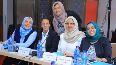 Fem kvinnor från Jemen med dukar på huvudet och plastflaskor framför sig sitter vid ett bord och ler mot kameran på en fredsförhandling .