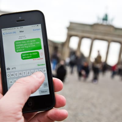 Ihminen pitää kännykkää kädessään niin, että ruudulla näkyy viestejä, taustalla Brandenburgin portti.