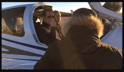 Två män sitter inne i styrhytten på ett litet flygplan. Framför dem står någon klädd i en svart rock med ryggen vänd mot kameran.