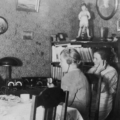 Svartvitt gammalt fotografi på barn och ungdomar som lyssnar på radio med hörlurar vid matbord