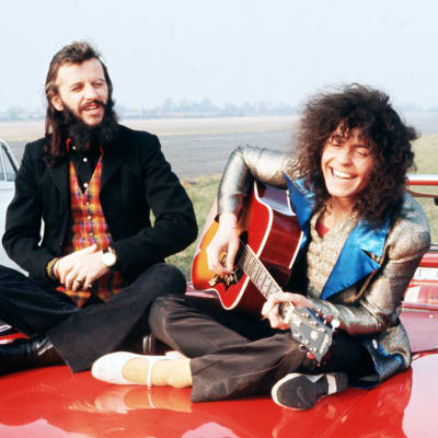 Ringo Starr och Marc Bolan sitter på motorhuv på ett fält. Bolan spelar akustisk gitarr och skrattar. Från filmen Born To Boogie 1972. Musiktestets logo.
