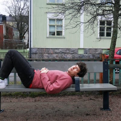 Kasper Strömman ligger på en parkbänk med fina trähus i bakgrunden i Trävallgård i Helsingfors.