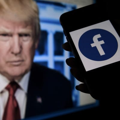 Puhelimen ruudulla näkyy Facebooking logo, taustalla näkyy Donald Trumpin kuva näytöllä.