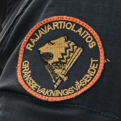 Anonym sjöbevakare i svart jacka. Gränsbevakningens emblem syns på axeln. En patrullbåt finns i bakgrunden.