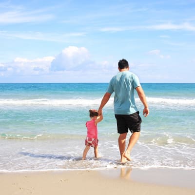 isä ja lapsi kävelevät käsikädessä rannalla vesirajassa