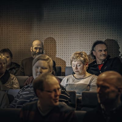 Folk sitter i ett auditorium. Ett par fotografer tar bilder av en kvinna.