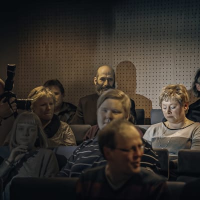 Folk sitter i ett auditorium. Ett par fotografer tar bilder av en kvinna.