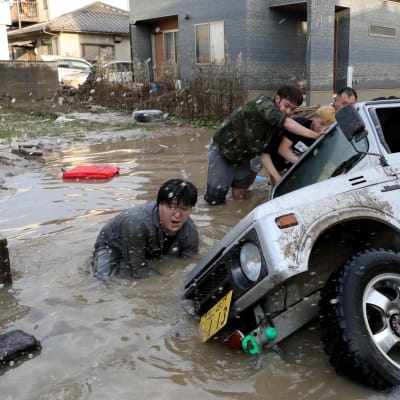 Miehet työntävät kaatunutta autoa pystyyn tulva-alueella Japanissa.