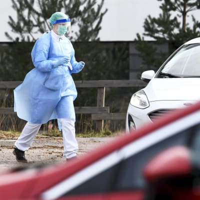 Vårdpersonal iklädd skyddsutrustning går mot vit bil för att ta ett drive-in coronatest av person.
