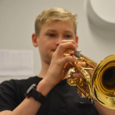 Närbild på en trumpet. I bakgrunden synns en pojke som spelar.