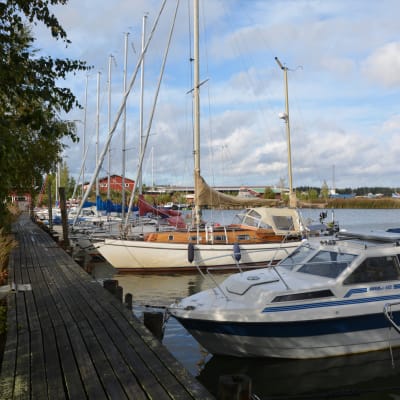 Flera småbåtar ligger förtöjda vid en brygga. I bakgrunden syns hamnbyggnader och båtar uppe på land.