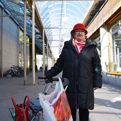 En äldre dam i svart jacka och röd hatt står bredvid en rollator. i stadsmiljö. Hon ler och tittar snett uppåt.