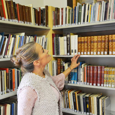 En blond kvinna står vid en bibliotekshylla fylld med böcker. Hon håller handen på en bokrygg och tittar uppåt mot böckerna ovanom henne.