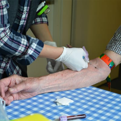 En hemvårdare tar blodprov av en äldre person. På bilden syns en bar arm och överkroppen av en person som håller på att ta blodprov med en nål.