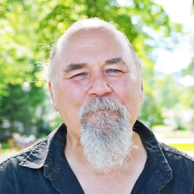 Närbild på en äldre man med grått skägg. Han står ute i en solig och grönskande park och ser glad ut.