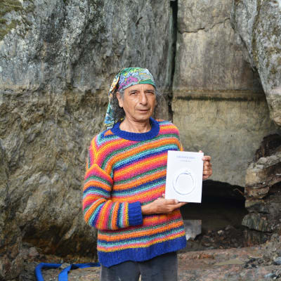 En medelålders man i randig tröja och duk om huvudet står framför en låg grottöppning. Han håller i en bok.