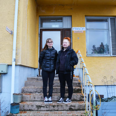 Två unga kvinnor i svarta ytterkläder står på en trappa utanför ett gult hus. På väggen finns en skylt där det står frisör.