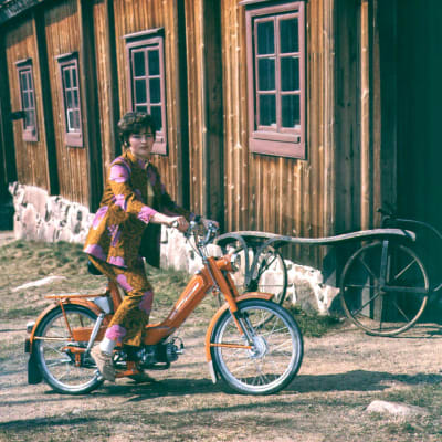Naistenvaatemainos kuvattuna Turun Luostarinmäellä,  nainen istuu mopon selässä kukallisessa housupuvussa.