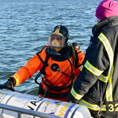 En dykare i orange dräkt vid vattenytan tillsammans med en annan person i svart jacka.